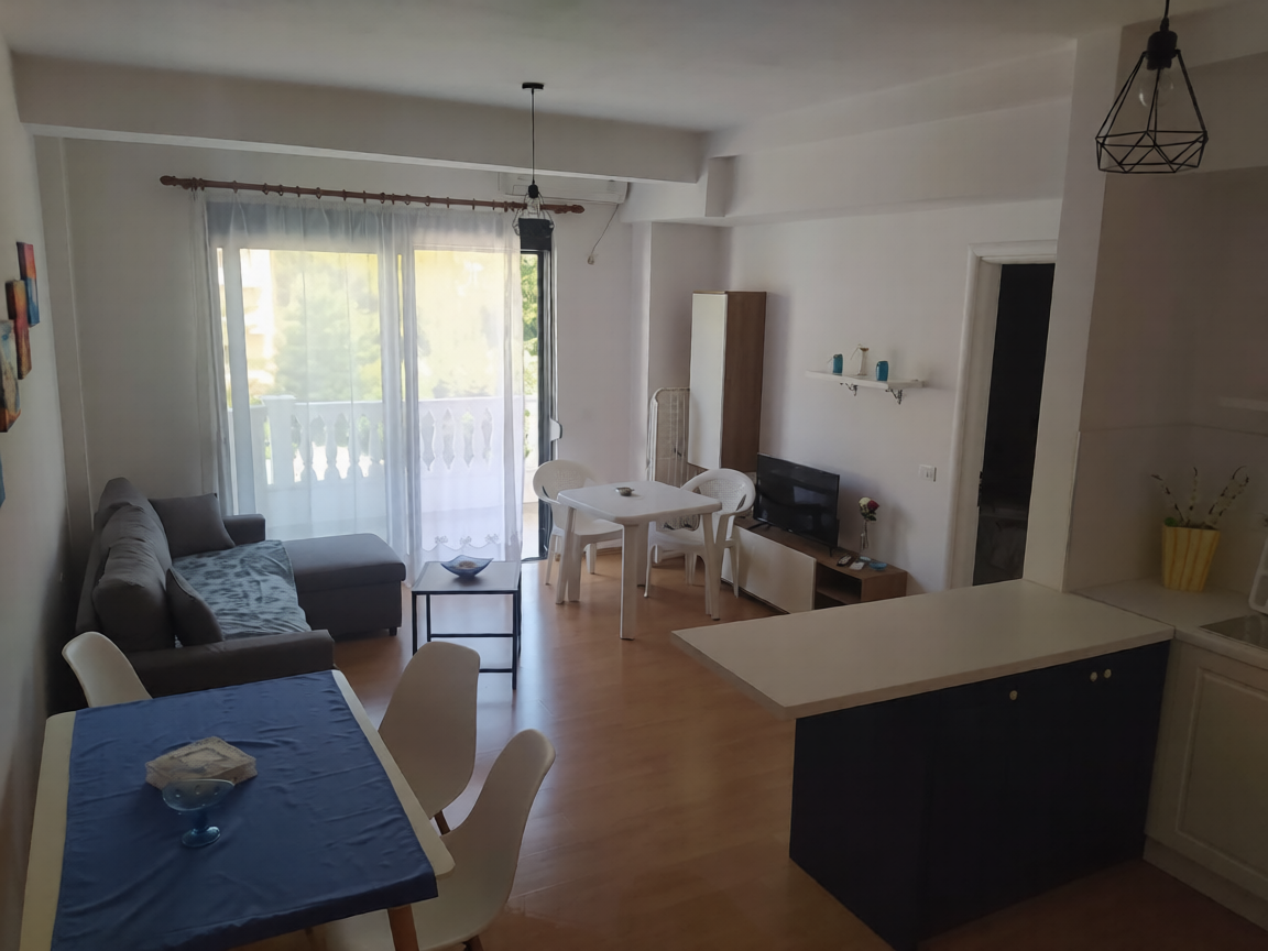 Modern apartment interior with open-plan living space, kitchen with black island, dining table with blue tablecloth, gray sofa, large windows with balcony, pendant light fixture